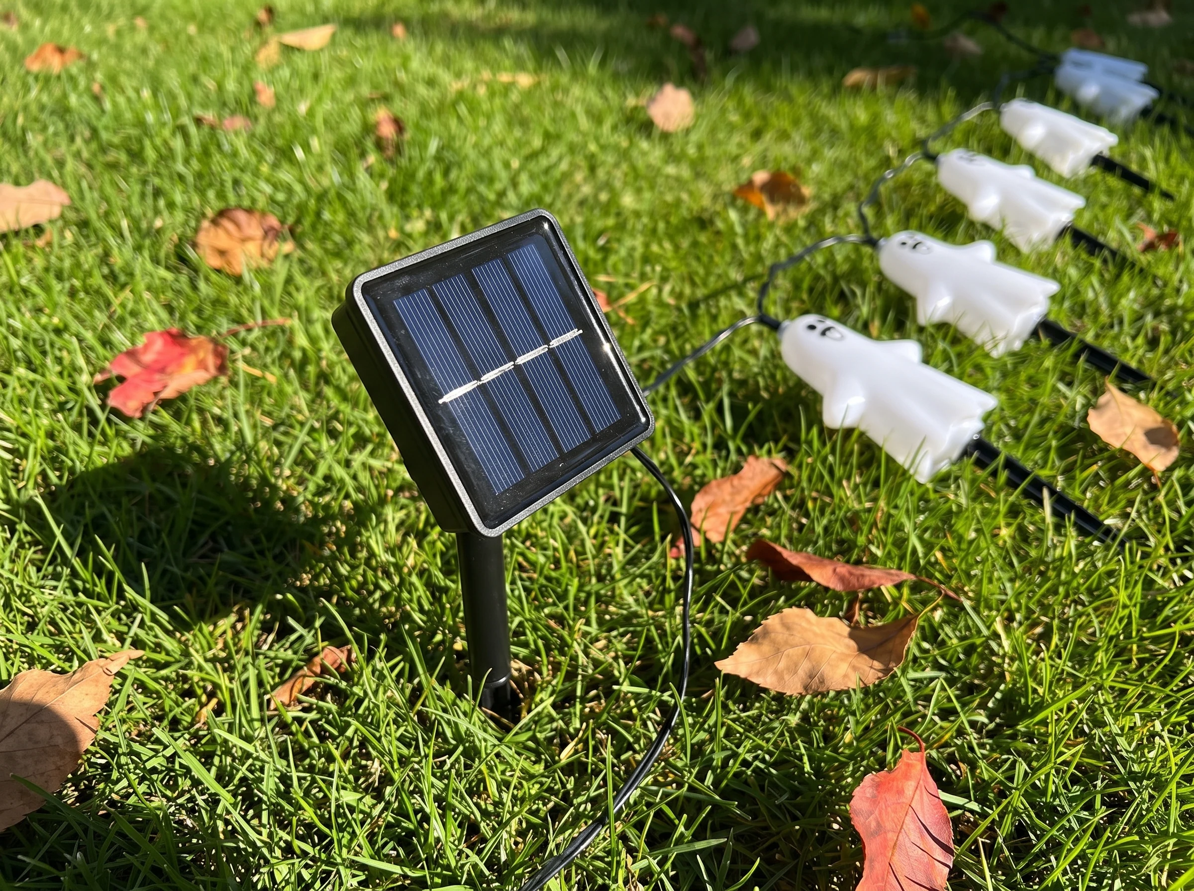 Close view of the small square solar panel on its own spike in the lawn, angled up to catch direct afternoon sun, with the wire running off to the ghosts