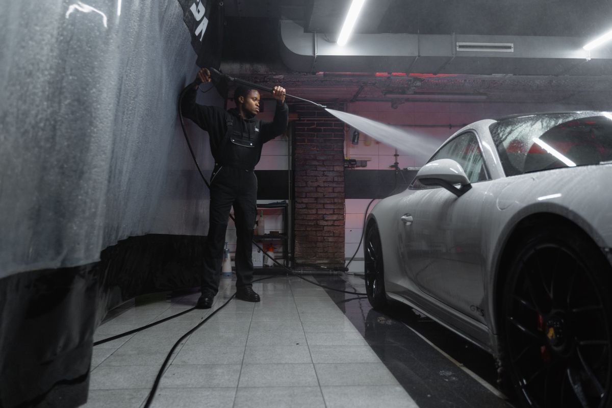 A man uses a pressure washer to clean a white car in an indoor car wash.