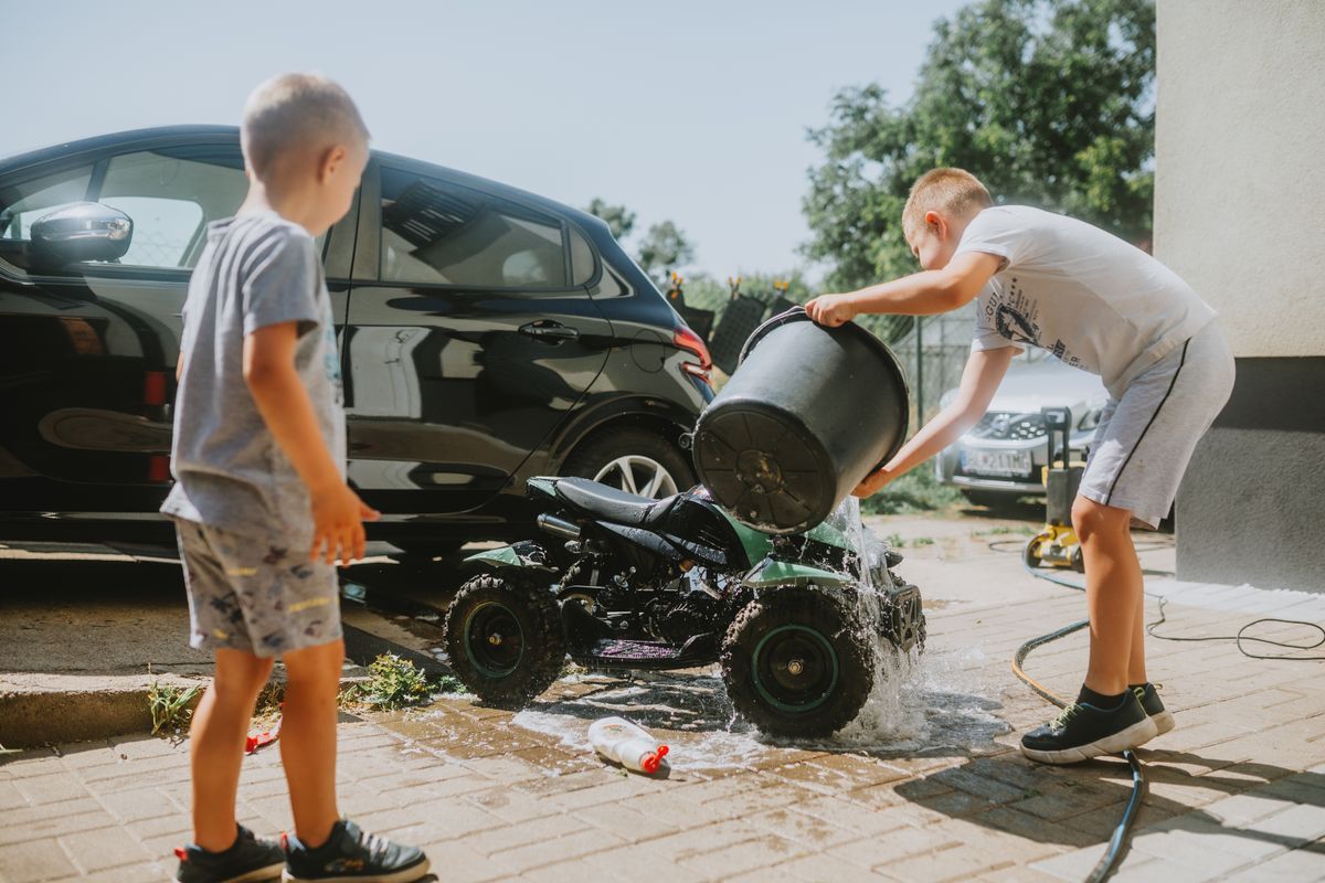 Two boys wash a toy ATV with a bucket near a car in a sunny driveway.