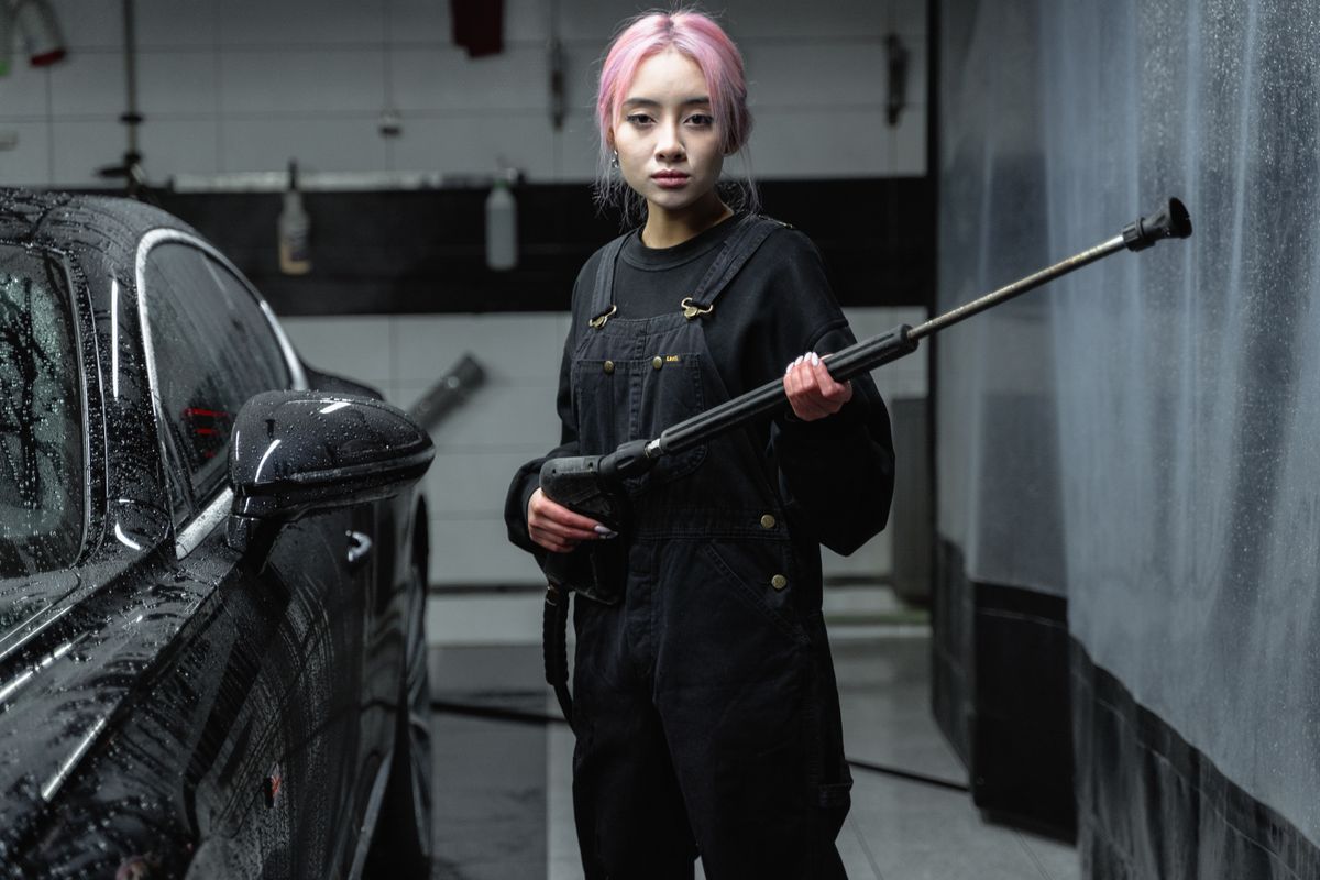 Asian woman with pink hair using a pressure washer at an indoor car wash.