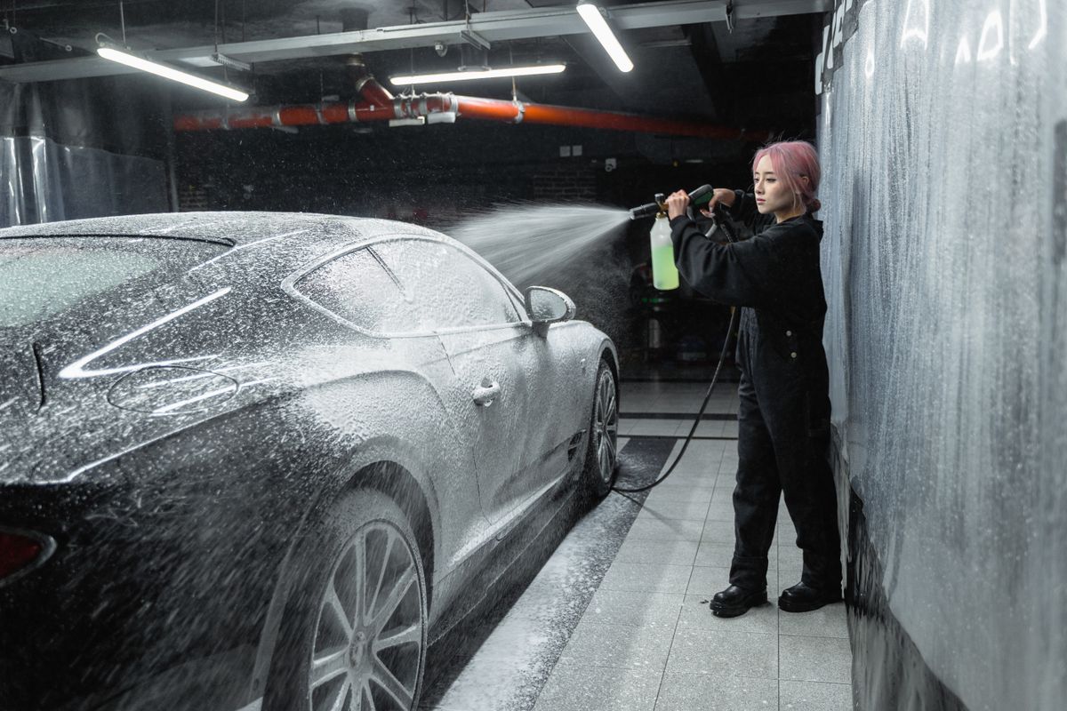 Asian woman with dyed hair washing a car indoors using a pressure washer.