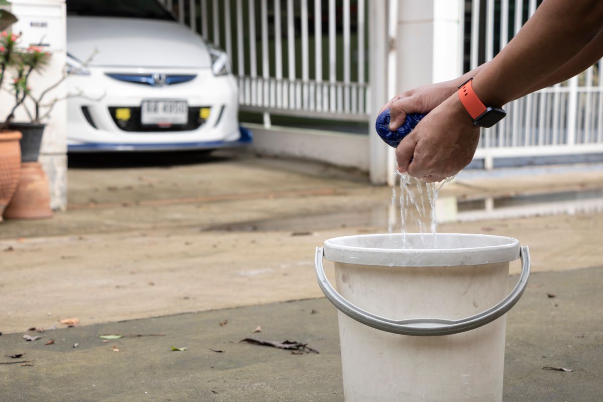 Man cleaning car with sponge and bucket in driveway, showcasing everyday car maintenance routine.