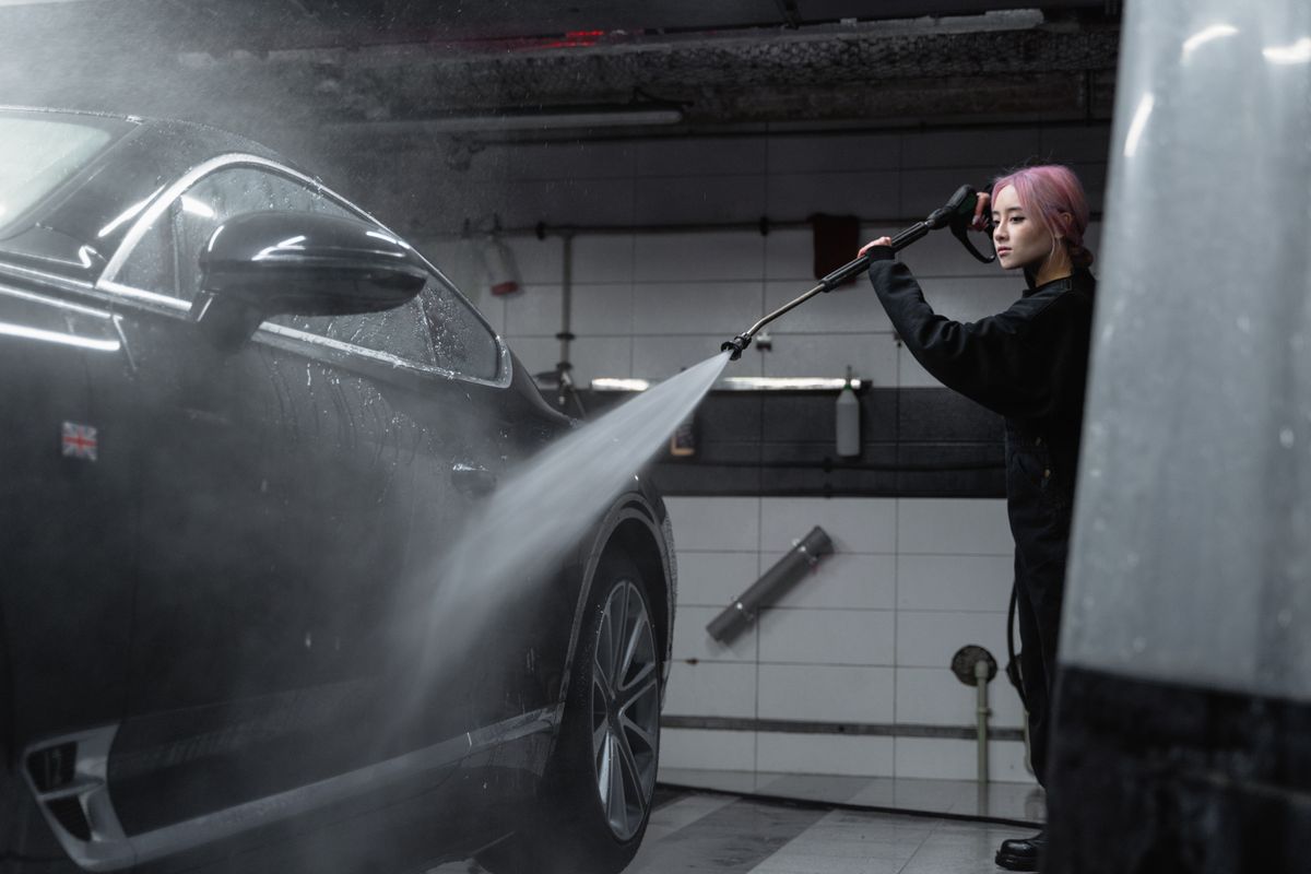 Woman power washing a black car in an indoor garage using a high-pressure washer.