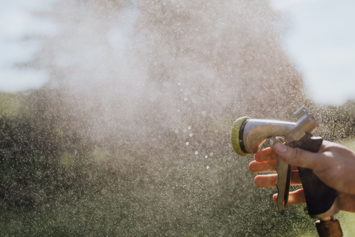 A hand holding a garden hose spray nozzle, misting water in an outdoor garden setting.