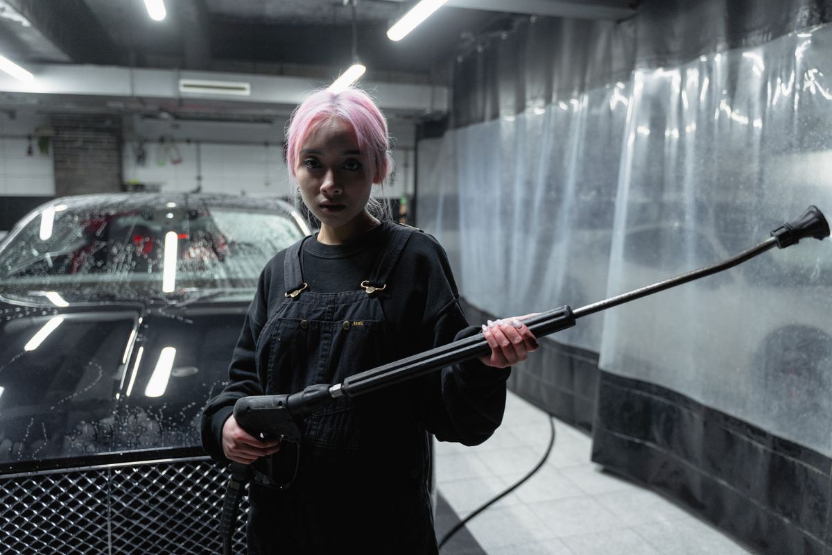 A woman with pink hair uses a pressure washer in a car wash setting. Indoor, nighttime shot.