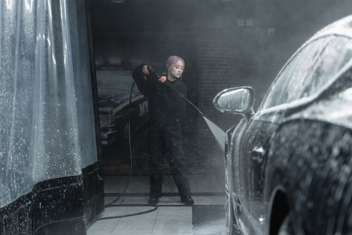 A young woman in a black outfit washing a car with foam and water spray indoors.