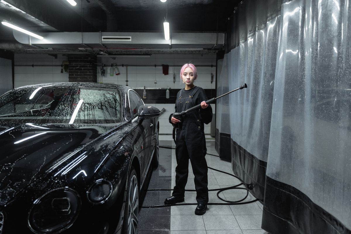 A woman using a pressure washer to clean a black sports car at an indoor car wash facility.