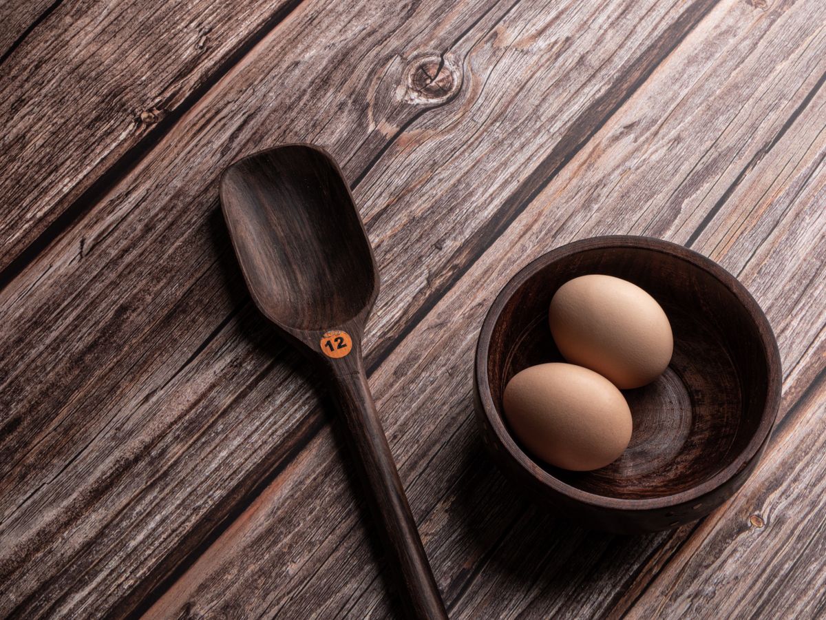 A rustic kitchen scene with a wooden bowl and eggs alongside a spoon on a wooden surface.