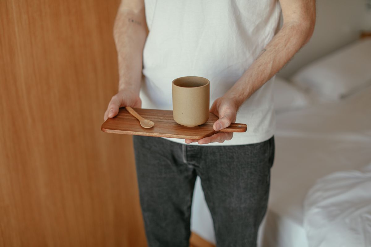 A man holding a wooden tray with a ceramic cup and spoon in a cozy indoor setting.