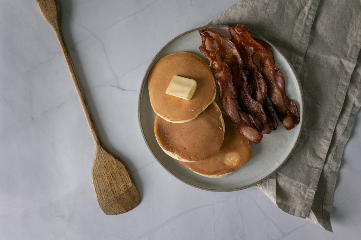 Top view of plate with tasty pancakes with butter near sliced roasted bacon near napkin and wooden kitchen spatula on white table in light kitchen