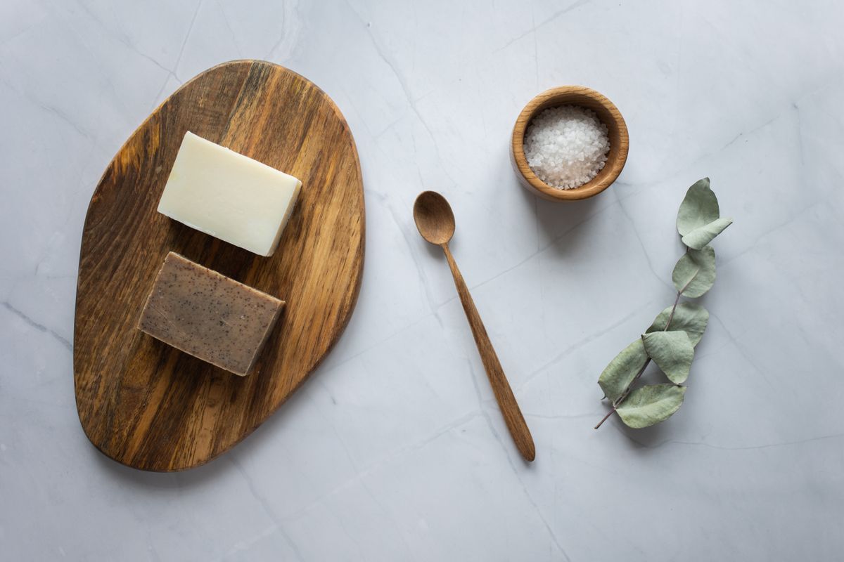 Top view of rectangular shaped soap on timber board composed with spoon and bowl with scrub near dry eucalyptus leaves