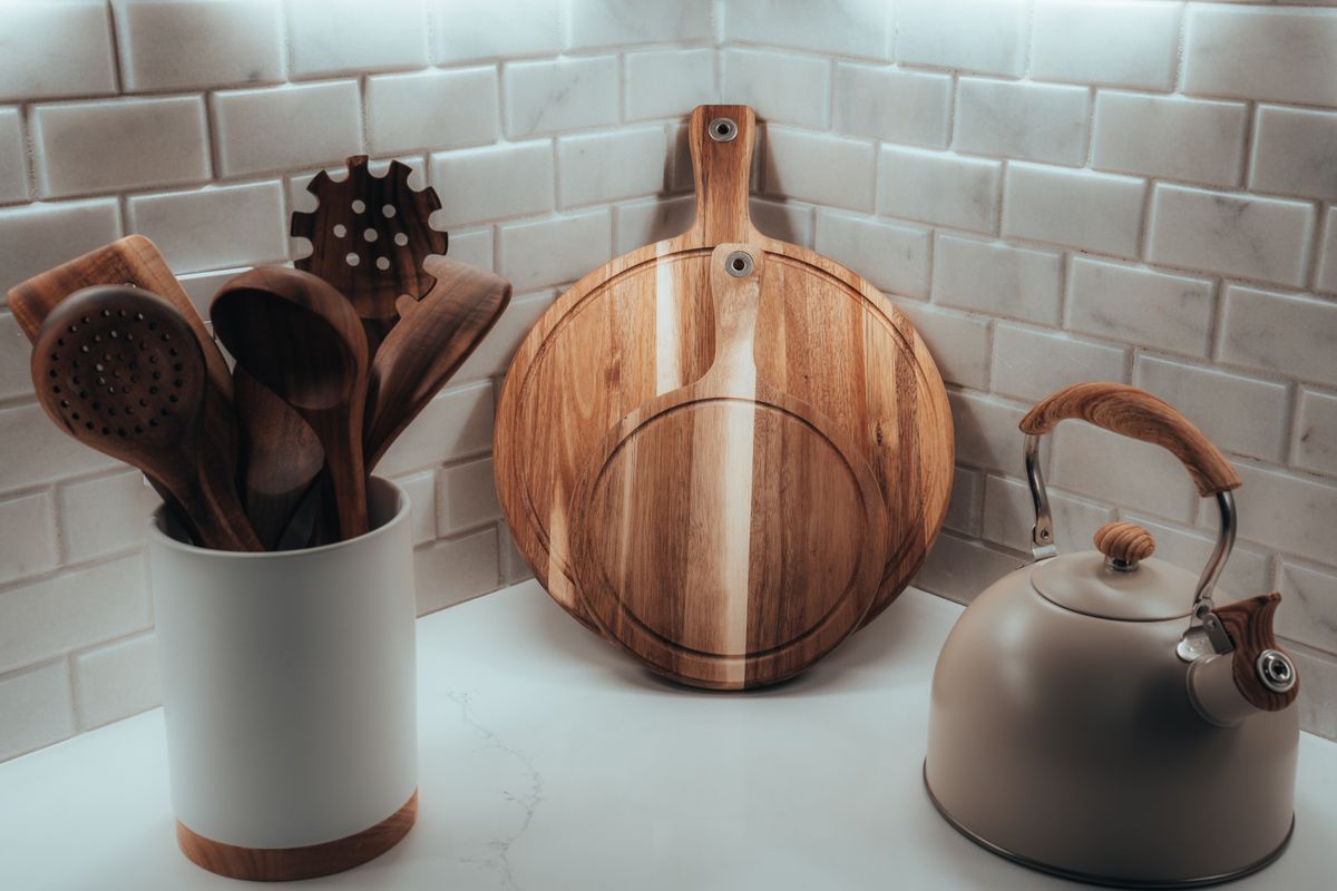 Cozy kitchen corner with wooden utensils, cutting board, and tea kettle against a tiled wall.