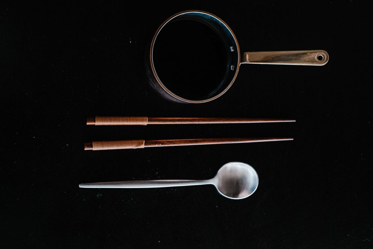 Flat lay of kitchen utensils including chopsticks, pot, and spoon on black surface.