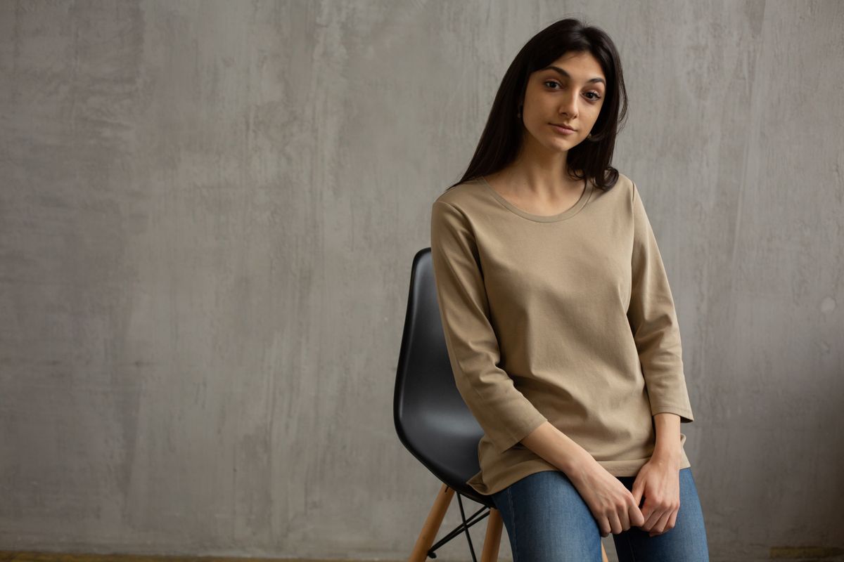 Young female wearing casual outfit sitting on chair and looking at camera against concrete wall