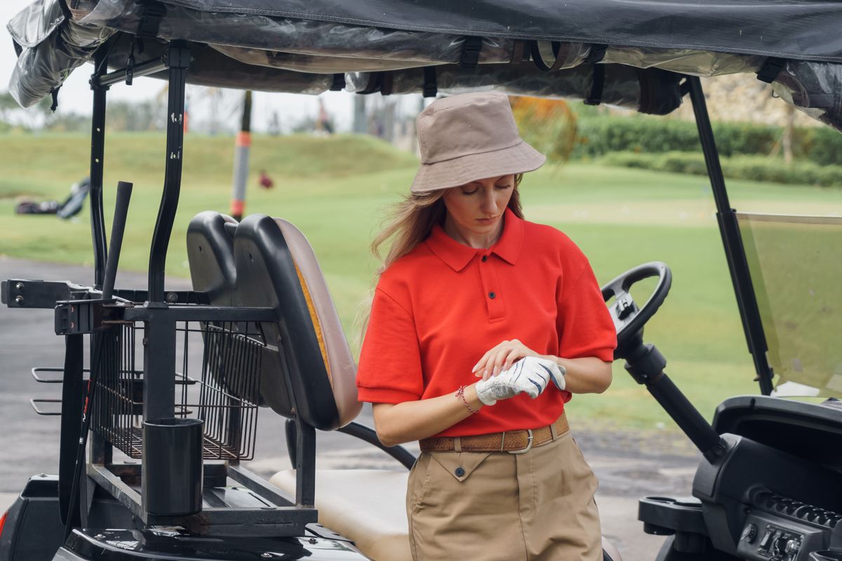 Stylish woman in red polo shirt and bucket hat preparing for golf game.