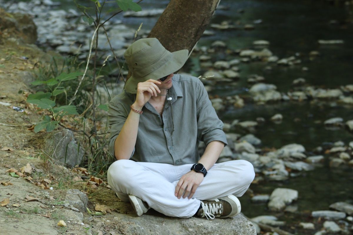 A man sitting calmly by a stream, wearing casual attire and a sun hat in a natural setting.