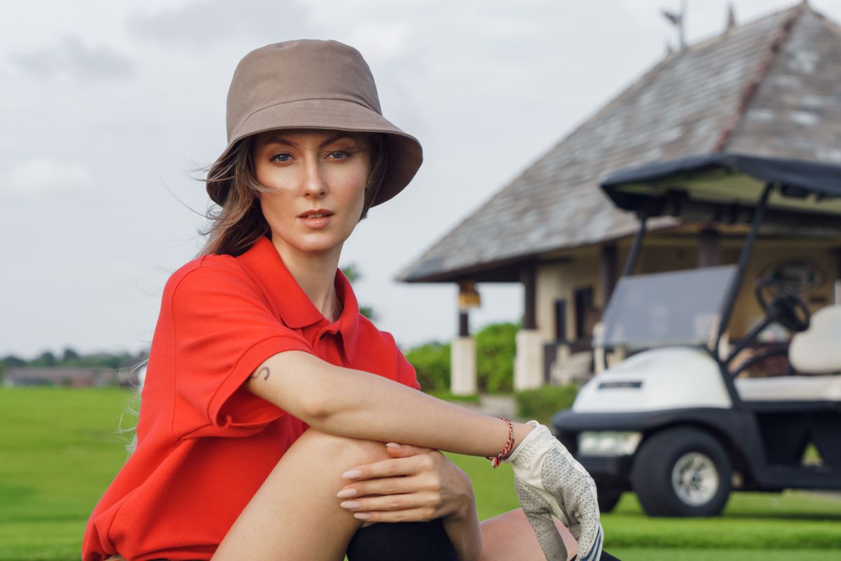 A woman in a red shirt and bucket hat sits on a golf course near a golf cart.
