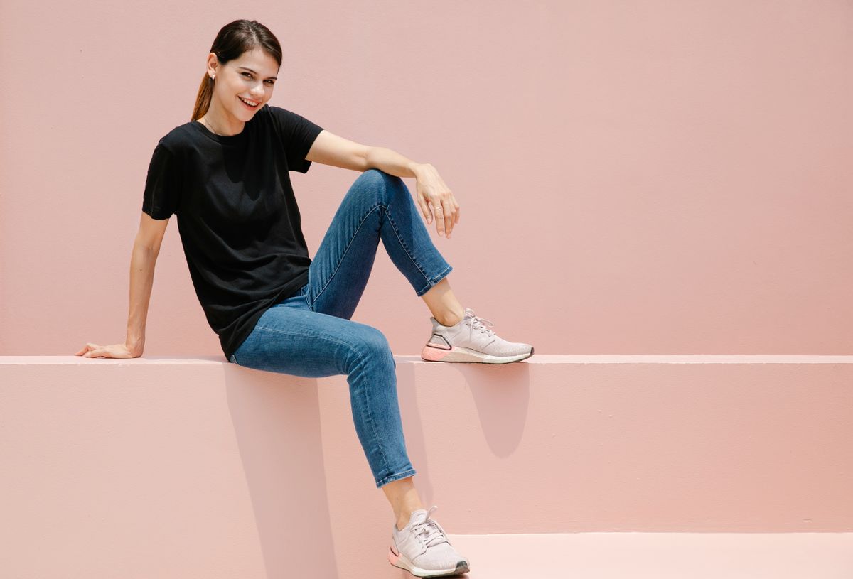 Young glad woman in t shirt and sneakers looking at camera while sitting on fence on pastel background