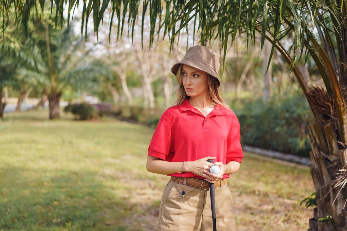 Woman golfer in red shirt with bucket hat holding a golf club outdoors.