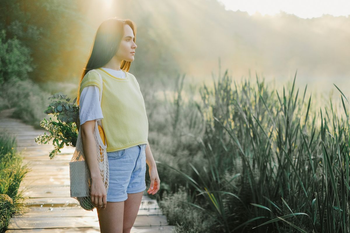 Woman wearing a yellow shirt and denim shorts standing on a sunny wooden pathway in nature.