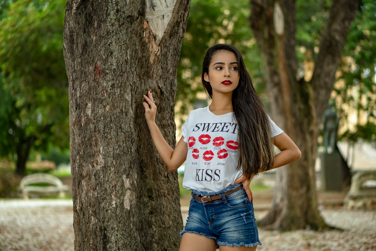 Serious girl with black long hair wearing denim shorts and t shirt standing near tree in green park