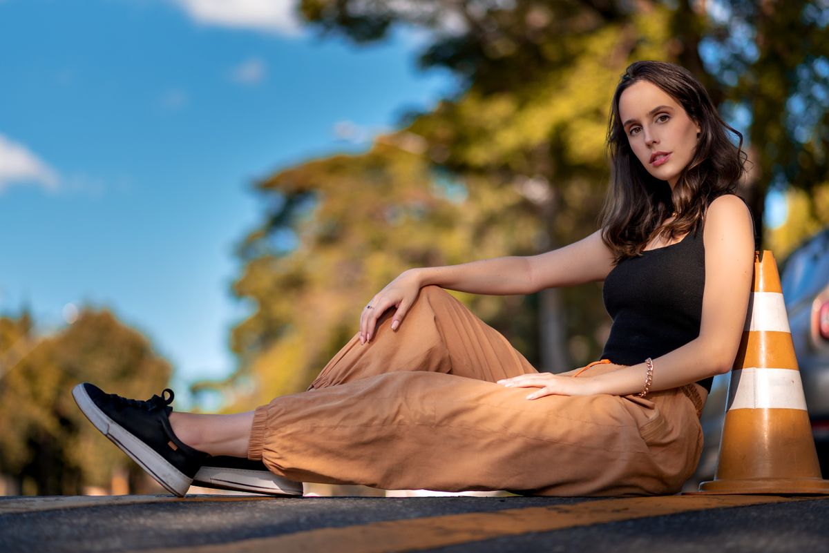 Stylish woman in black top and brown pants poses creatively next to a traffic cone in an outdoor urban setting.