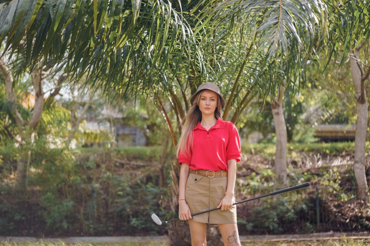 Woman golfer in red polo shirt and hat standing with a golf club under palm trees.