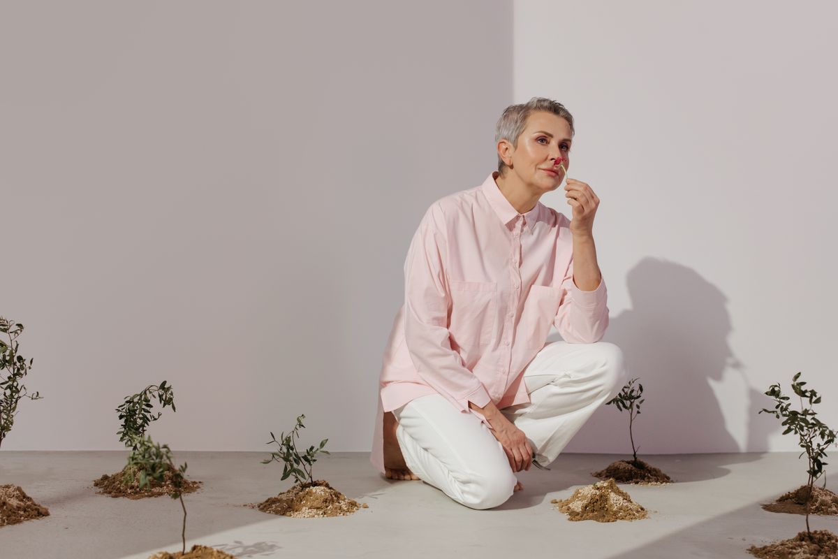 Senior woman crouching among small plants in a minimalist indoor setting, wearing a pink shirt and white pants.