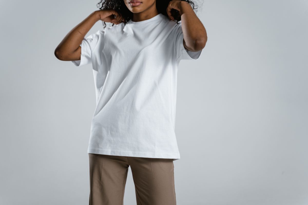 Woman wearing a white t-shirt and brown pants, adjusting her hair against a neutral background.