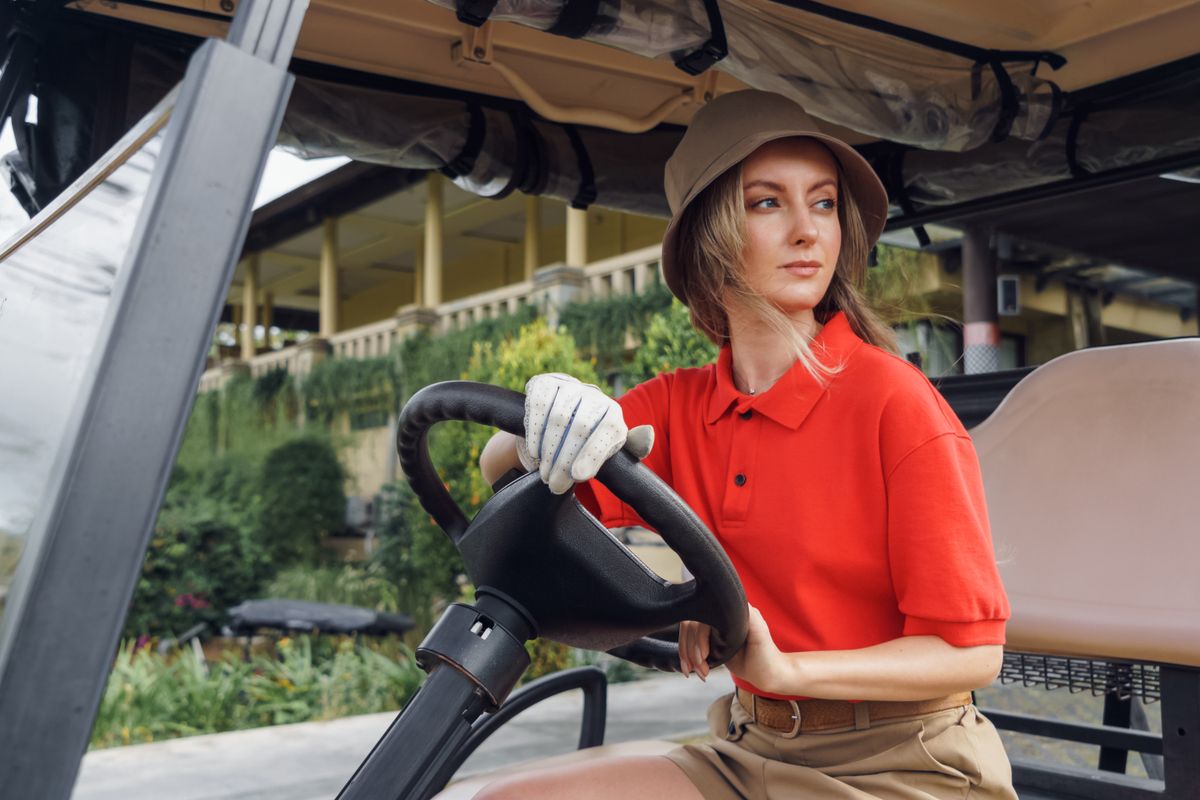 Woman wearing red shirt and bucket hat driving a golf cart, looking sideways.