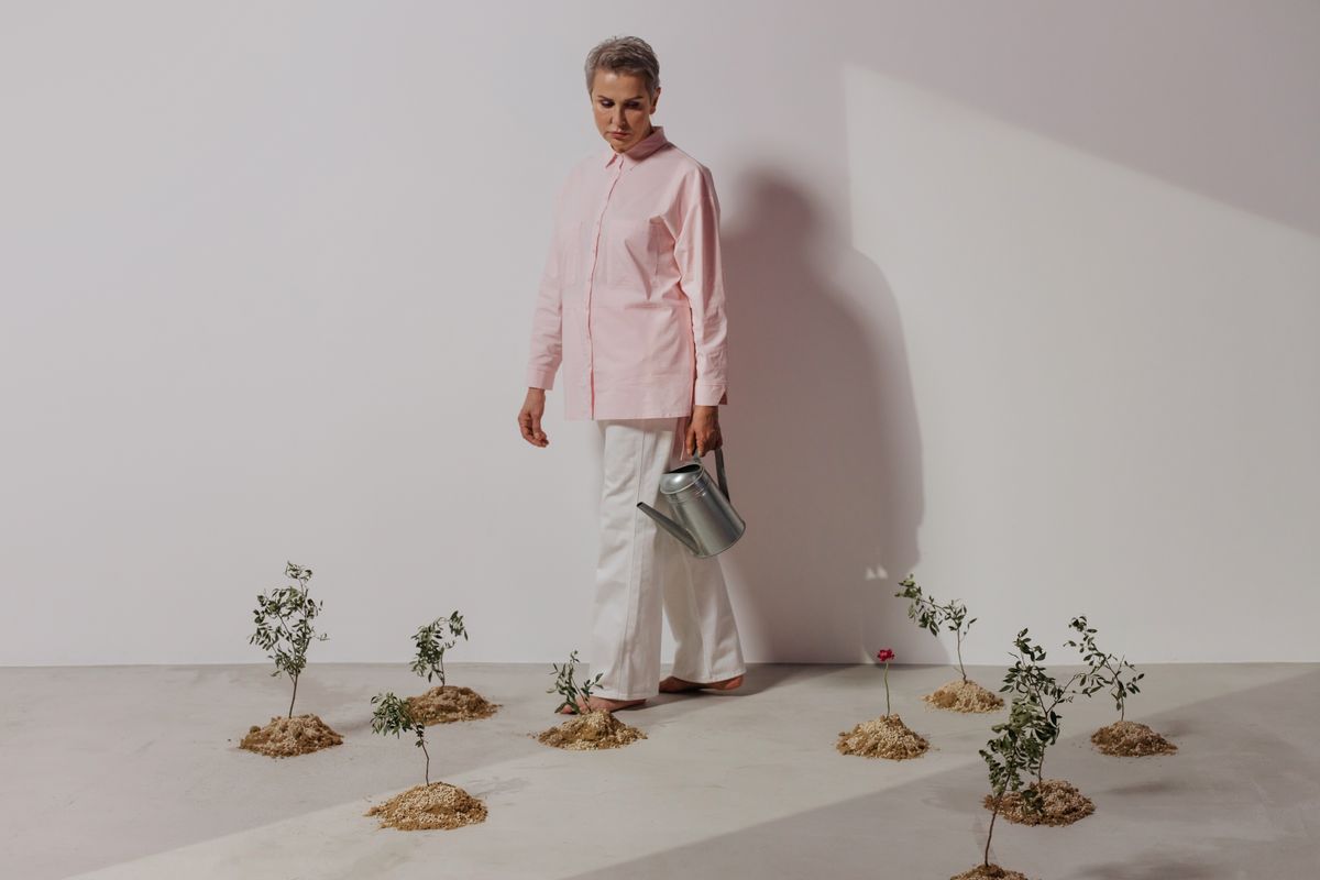 Elegant woman wearing a pink shirt, holding a watering can, standing among plants.