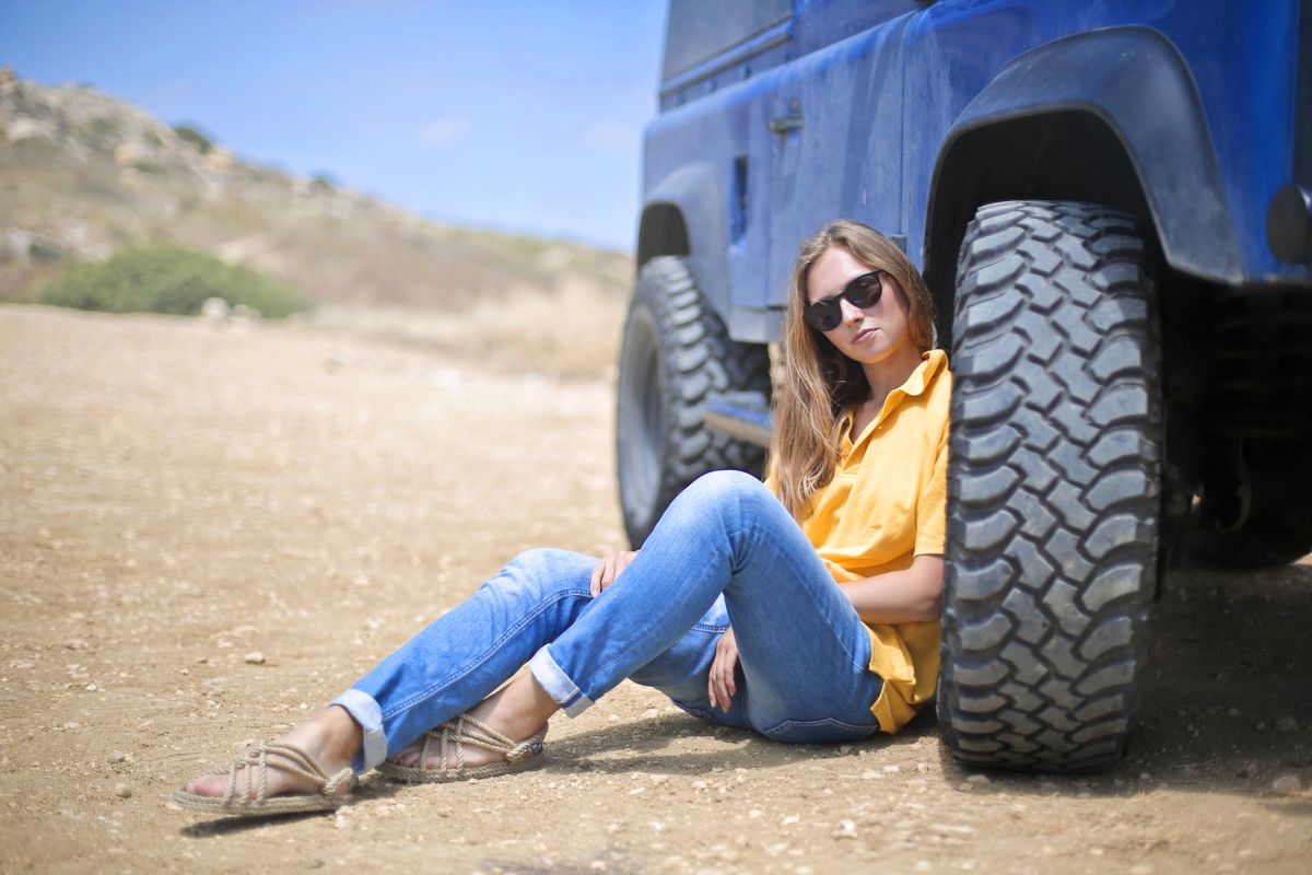 A woman wearing sunglasses relaxes against a blue off-road vehicle in a desert landscape.