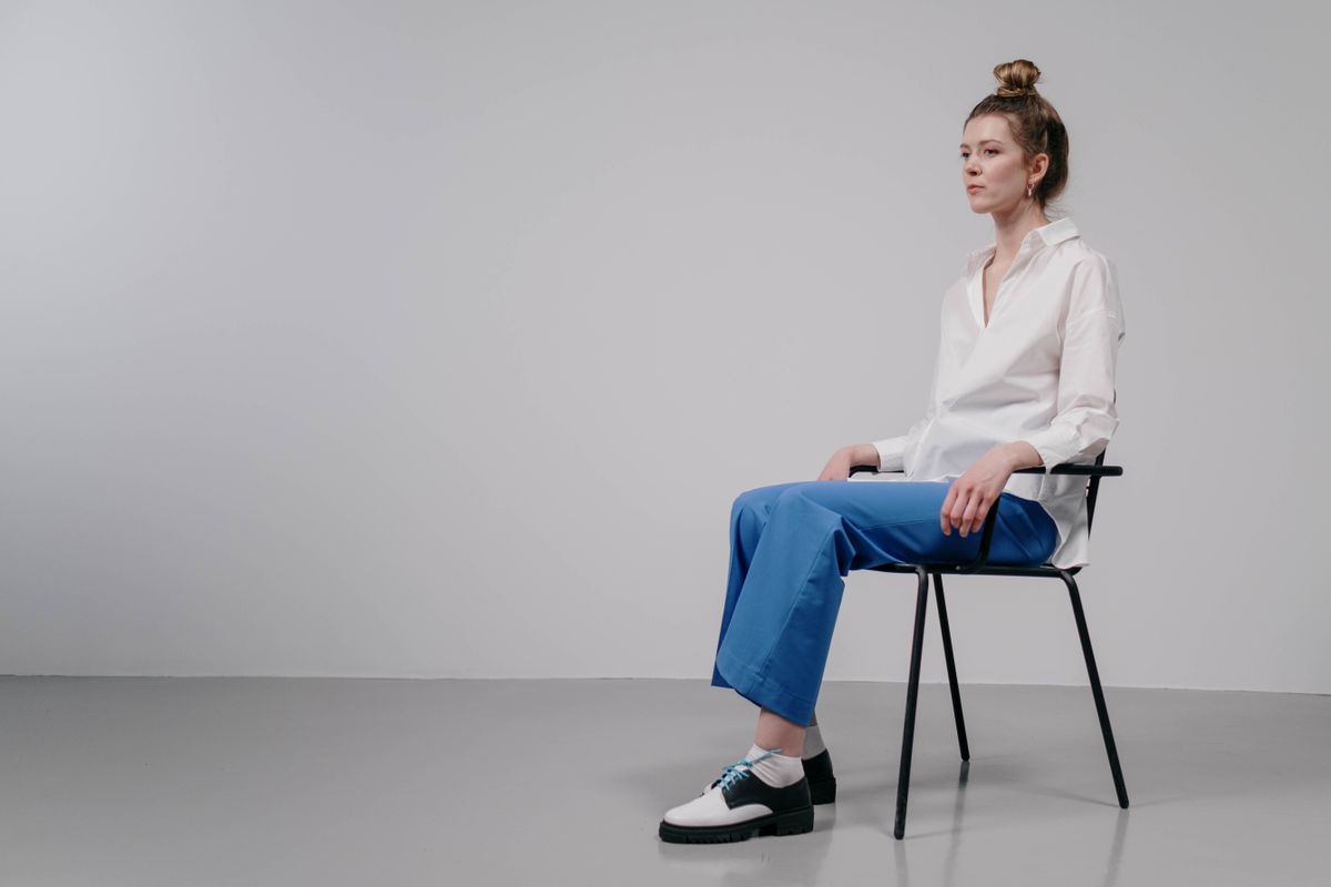 A woman in a white shirt and blue pants sits on a chair in a minimalist studio setting.