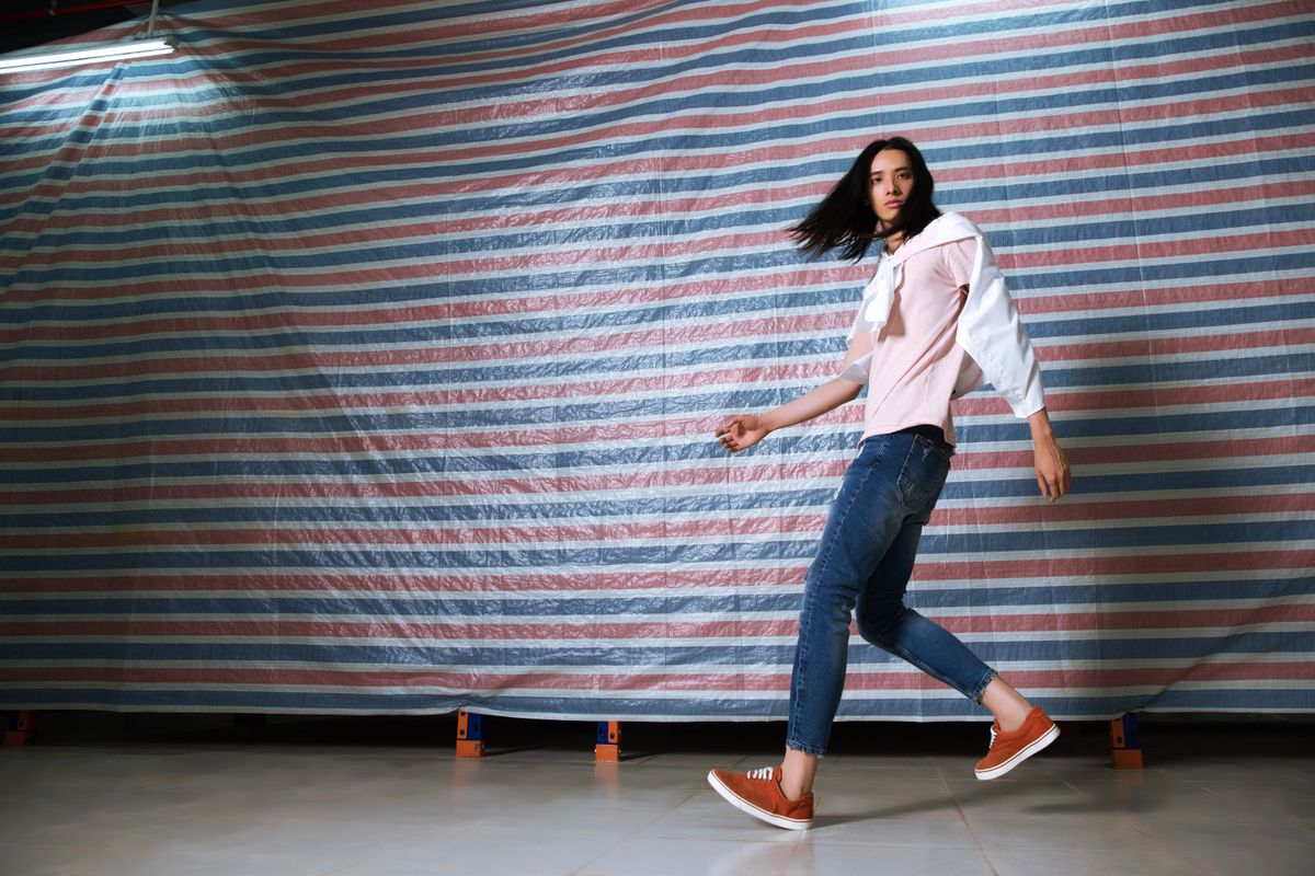 Fashion model posing indoors with a casual style in denim and sneakers against a striped background.