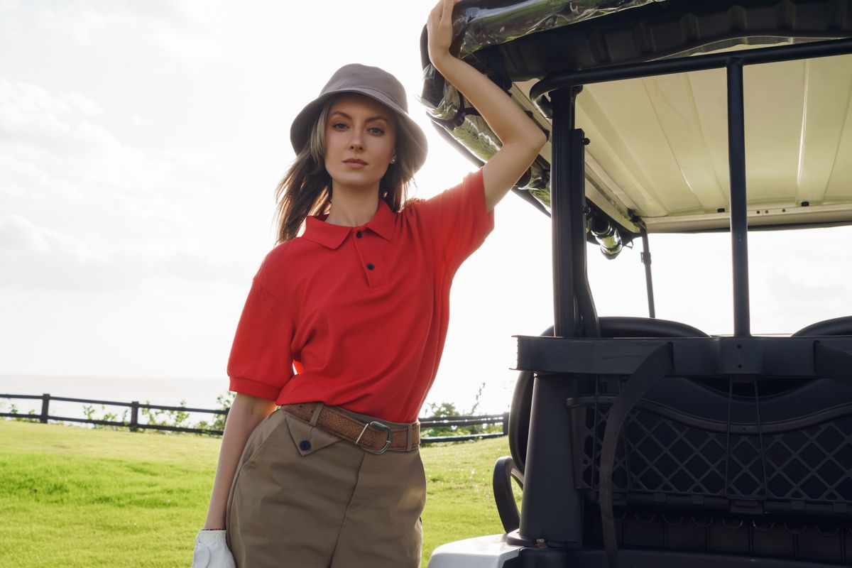 Woman in red shirt and bucket hat stands by a golf cart on a sunny golf course.