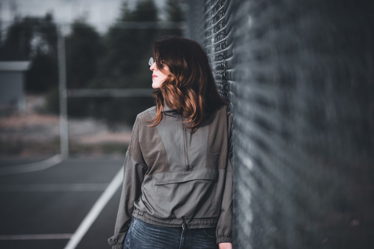 A fashionable woman in a casual jacket poses thoughtfully outdoors by a chain link fence.