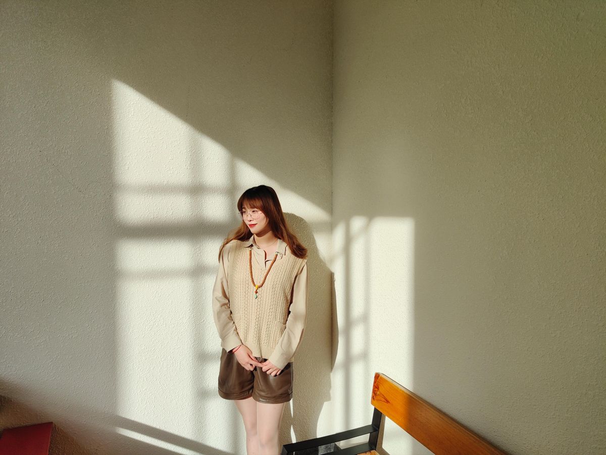 A young woman stands in a sunlit corner, wearing a beige sweater vest, creating a warm and serene atmosphere.