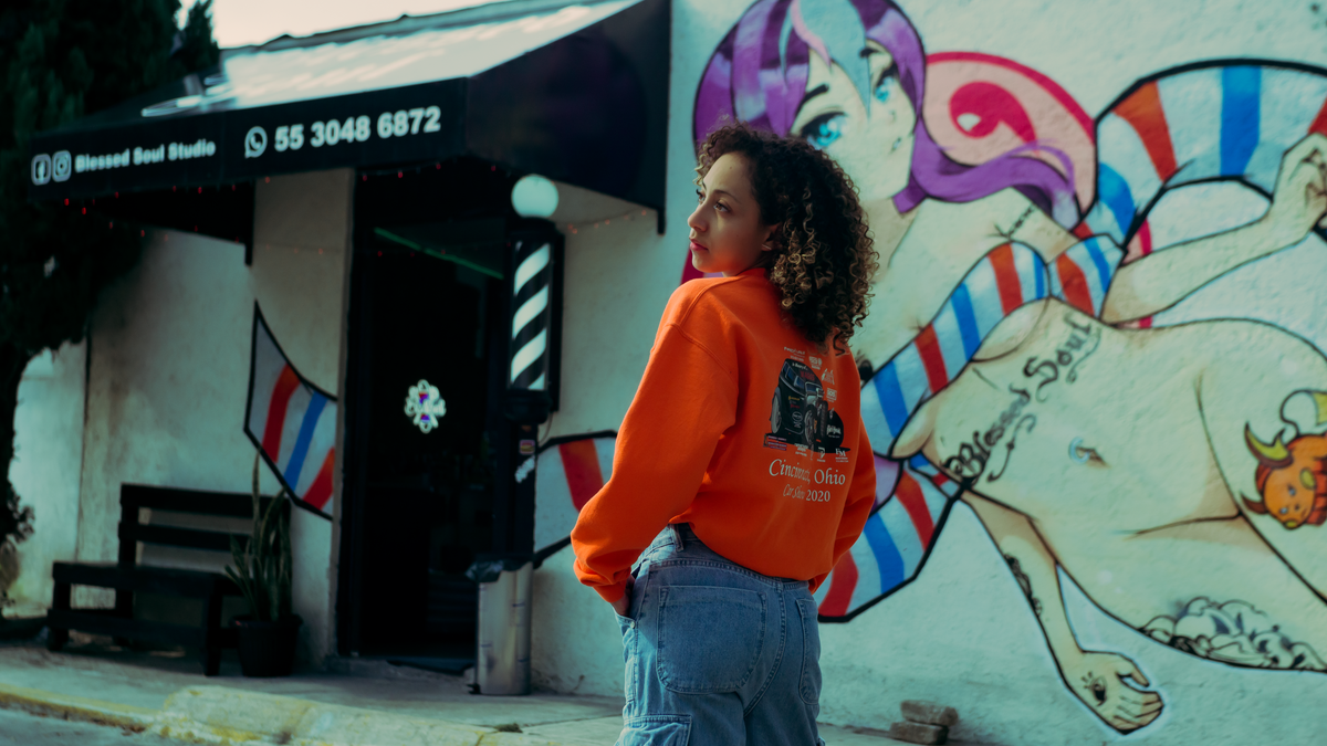 Young woman in vibrant outfit poses in front of urban graffiti wall near barbershop.