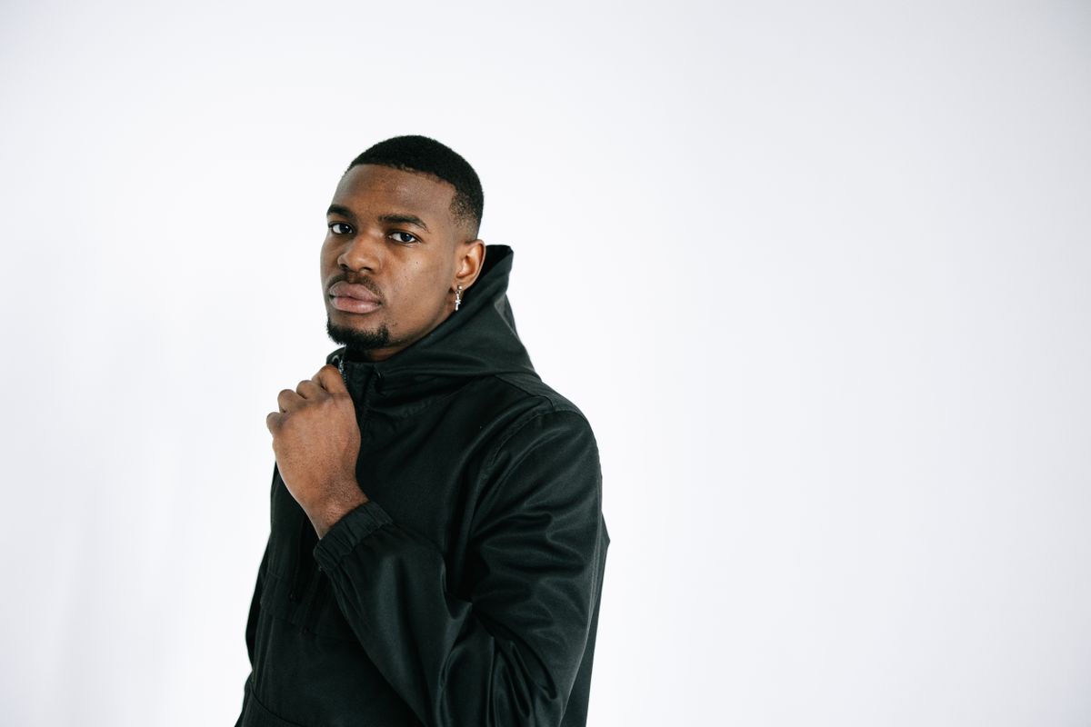 A close-up portrait of a serious man wearing a black hoodie against a white background.