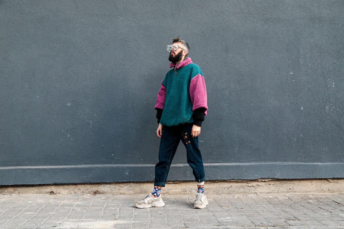 Stylish man wearing colorful sweater and jeans stands by gray wall.