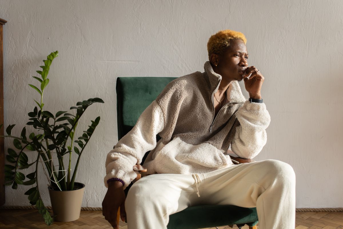 Stylish young man with blonde hair in a fur coat sitting indoors, deep in thought.