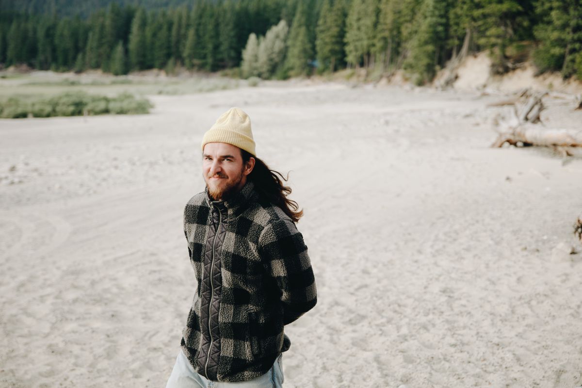 Bearded man in checkered jacket and beanie enjoying a calm walk on a sandy path outdoors.