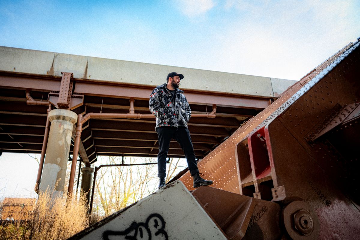Young man standing under an industrial bridge, showcasing urban fashion style in a rugged environment.