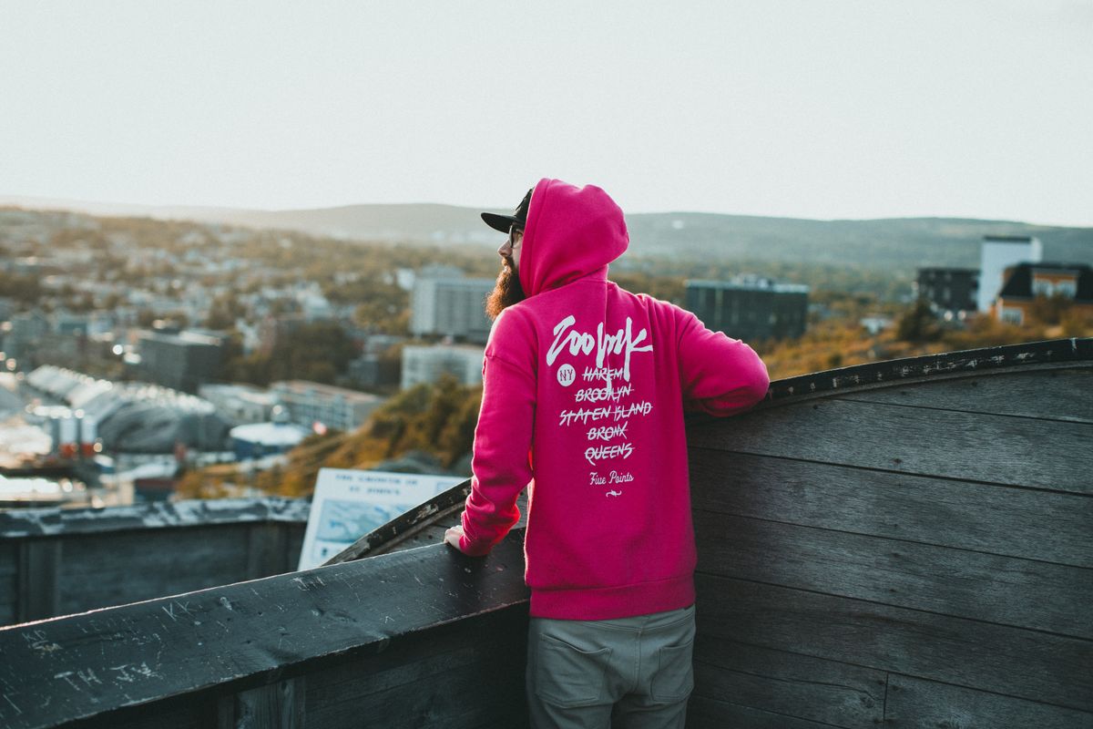 Man wearing a vibrant pink hoodie gazing at cityscape from a wooden deck in daylight.