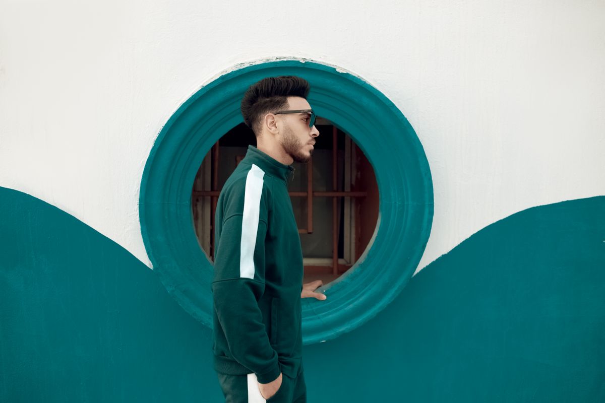 Stylish young man stands confidently in front of a unique circular window, showcasing modern fashion.