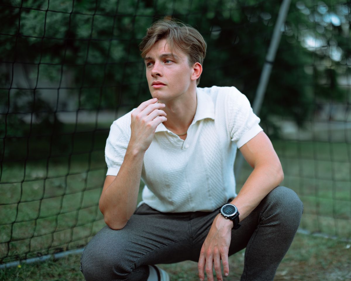 Young man with thoughtful pose in a white shirt outdoors, near a garden fence.