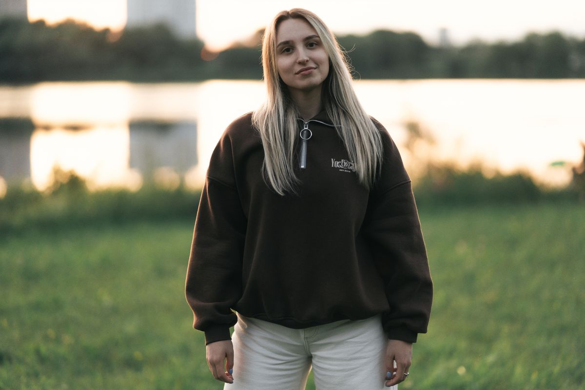 Portrait of a young woman in a brown hoodie and white pants standing by a lake at sunset.