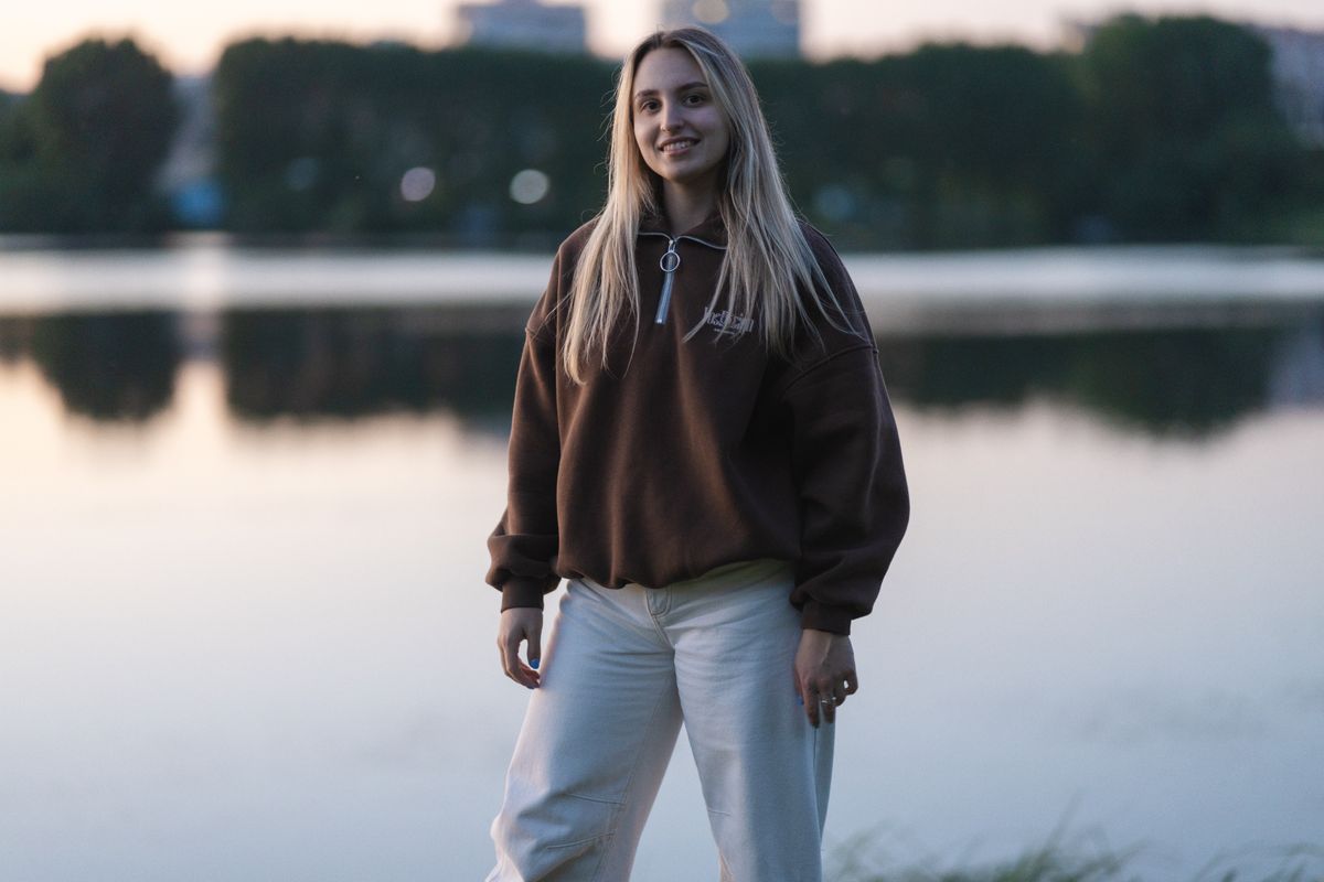 A young woman stands by a calm lake at twilight wearing a brown sweater and white pants, smiling confidently.