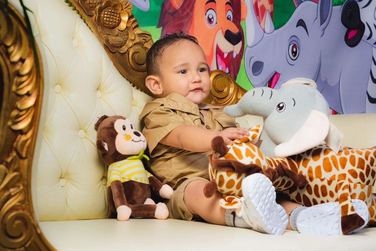 Charming toddler playing with plush toys on an ornate chair against a colorful backdrop.