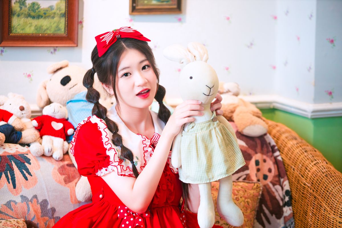 Young girl in a vibrant red dress indoors, holding a plush rabbit toy surrounded by stuffed animals.